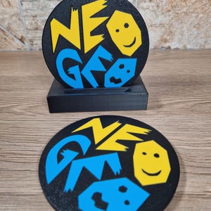 May include: Black round coaster with the word "NEO GEO" in yellow and blue. The coaster has a smiling face graphic. The coaster is sitting on a black stand. The coaster is made of a hard material.
