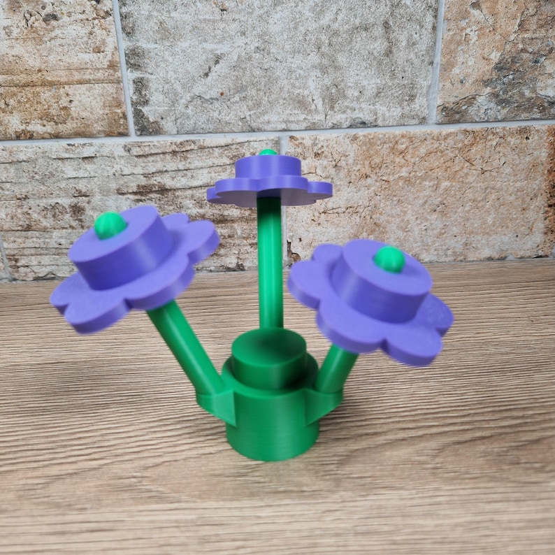 May include: A 3D-printed flower sculpture featuring three purple flowers with green centers and stems. The base and connecting elements are green. The sculpture is displayed on a wooden surface with a brick background.