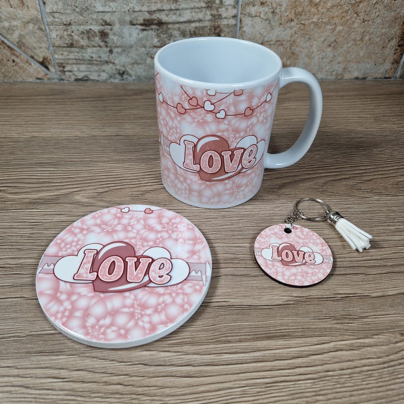 May include: A white ceramic mug, coaster, and keychain set. The items feature a pink floral pattern with the word "Love" in a heart-shaped design. The keychain has a silver ring and a white tassel.