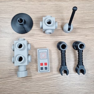 May include: An assortment of gray and black plastic toy parts, including a circular base, several cube-shaped connectors, a lever, a control panel with red and black squares, and two claw-like pieces. The parts are arranged on a wooden surface.