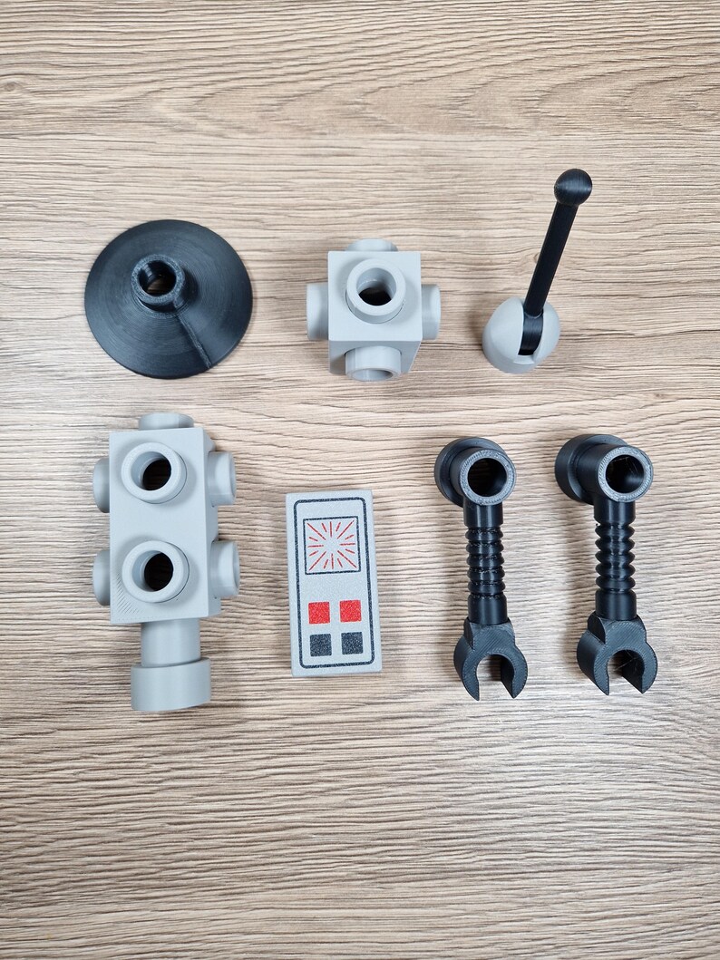 May include: An assortment of gray and black plastic toy parts, including a circular base, several cube-shaped connectors, a lever, a control panel with red and black squares, and two claw-like pieces. The parts are arranged on a wooden surface.