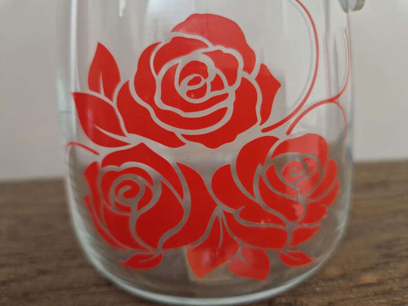 May include: A clear glass jar with a red rose design. The roses are outlined in white and have a vintage look.