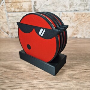 May include: A set of red coasters with a black border and a black sunglasses design. The coasters are stacked on a black rectangular holder. The design features a black dot and white accents on the sunglasses.