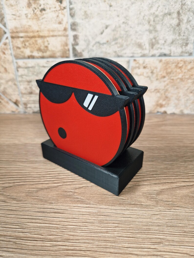 May include: A set of red coasters with a black border and a black sunglasses design. The coasters are stacked on a black rectangular holder. The design features a black dot and white accents on the sunglasses.