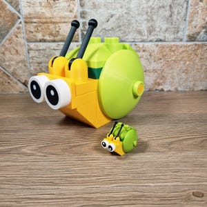 Puede incluir: Un juguete de caracol amarillo y verde hecho de bloques entrelazados, con grandes ojos con borde blanco y antenas negras. Tambi&eacute;n hay un juguete de insecto m&aacute;s peque&ntilde;o a juego. Los juguetes est&aacute;n sobre una superficie de madera.