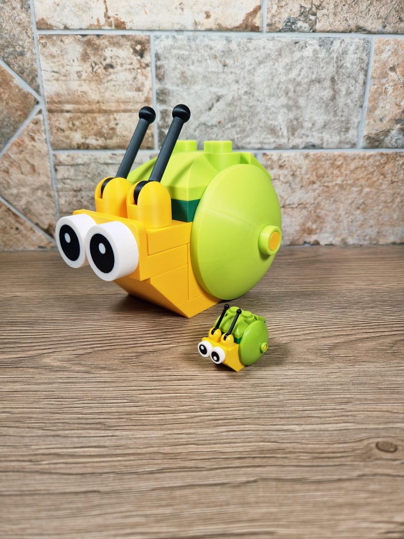 Puede incluir: Un juguete de caracol amarillo y verde hecho de bloques entrelazados, con grandes ojos con borde blanco y antenas negras. Tambi&eacute;n hay un juguete de insecto m&aacute;s peque&ntilde;o a juego. Los juguetes est&aacute;n sobre una superficie de madera.