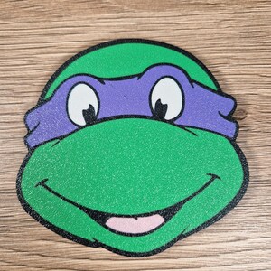 Puede incluir: Primer plano de la cara de una Tortuga Ninja, verde y morada, con una boca sonriente, ojos blancos y un contorno negro. La imagen representa la cara del personaje.