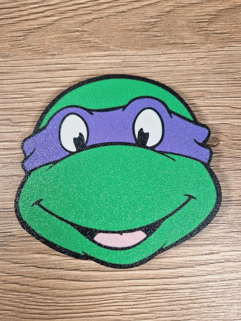 Puede incluir: Primer plano de la cara de una Tortuga Ninja, verde y morada, con una boca sonriente, ojos blancos y un contorno negro. La imagen representa la cara del personaje.