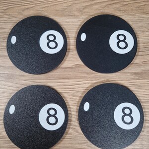 May include: Four black round coasters with a textured surface, each featuring a white number 8 and a white dot. The coasters are arranged in a square formation on a light brown wooden surface.
