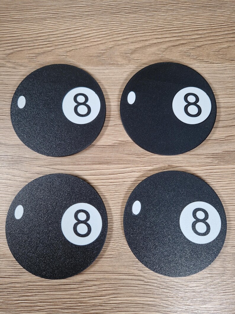 May include: Four black round coasters with a textured surface, each featuring a white number 8 and a white dot. The coasters are arranged in a square formation on a light brown wooden surface.