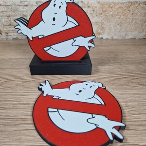 K&ouml;nnte beinhalten: Zwei Ghostbusters-Logo-Dekorationen. Eine ist eine Standfigur auf einem schwarzen Sockel, die andere ein flacher Untersetzer. Beide zeigen den ikonischen roten Kreis mit einem Geist und einem Schr&auml;gstrich.
