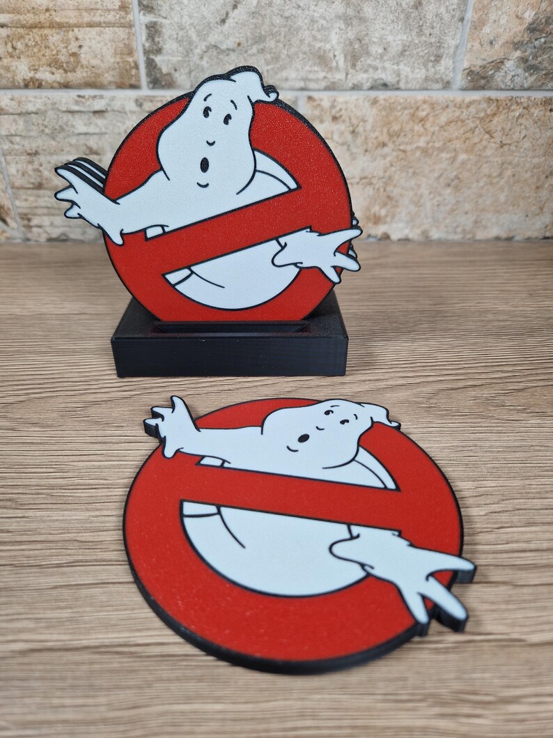 K&ouml;nnte beinhalten: Zwei Ghostbusters-Logo-Dekorationen. Eine ist eine Standfigur auf einem schwarzen Sockel, die andere ein flacher Untersetzer. Beide zeigen den ikonischen roten Kreis mit einem Geist und einem Schr&auml;gstrich.