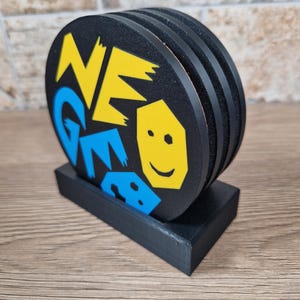 May include: A set of black coasters with a matching holder. Each coaster has a unique design with the word "NEO GEO" in yellow and blue, along with a yellow smiling face. The coasters are stacked and displayed in a black holder.