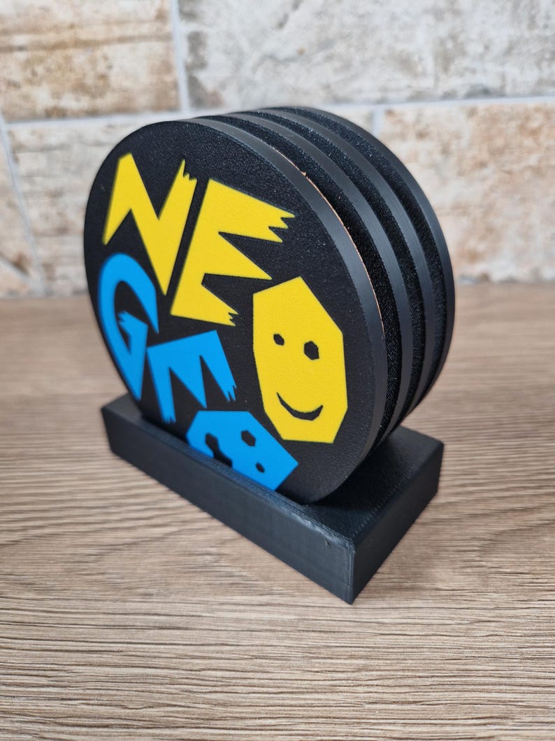May include: A set of black coasters with a matching holder. Each coaster has a unique design with the word "NEO GEO" in yellow and blue, along with a yellow smiling face. The coasters are stacked and displayed in a black holder.