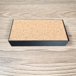 May include: A rectangular cork coaster with a black border. The coaster has a natural cork surface and a black painted edge. The coaster is sitting on a light brown wooden surface.