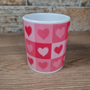 May include: A white ceramic mug with a pink and red heart pattern. The design features a checkerboard pattern of pink squares, each containing a red or pink heart. The mug is cylindrical and sits on a wooden surface.