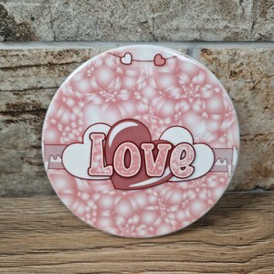 May include: A round, pink coaster with a bubble-like pattern. The word "Love" is displayed in a heart-shaped design, with a pink heart and white clouds. Two small heart shapes are at the top.