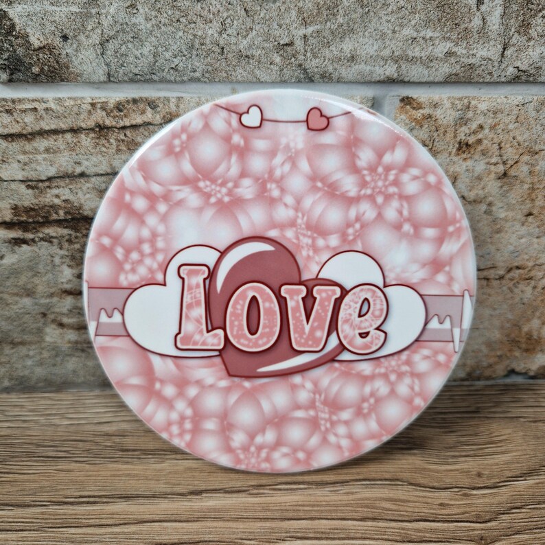 May include: A round, pink coaster with a bubble-like pattern. The word "Love" is displayed in a heart-shaped design, with a pink heart and white clouds. Two small heart shapes are at the top.