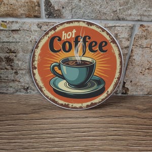 May include: Round metal sign with a vintage design featuring a teal coffee cup on a saucer, with steam rising from the coffee. The words "hot Coffee" are in a stylized font against an orange background with a distressed border.