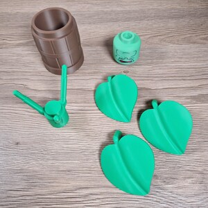 May include: An assortment of 3D-printed items, including a brown barrel-shaped container, a green head-shaped object with a face, a green tool, and three green leaf-shaped objects. The items are arranged on a wooden surface.