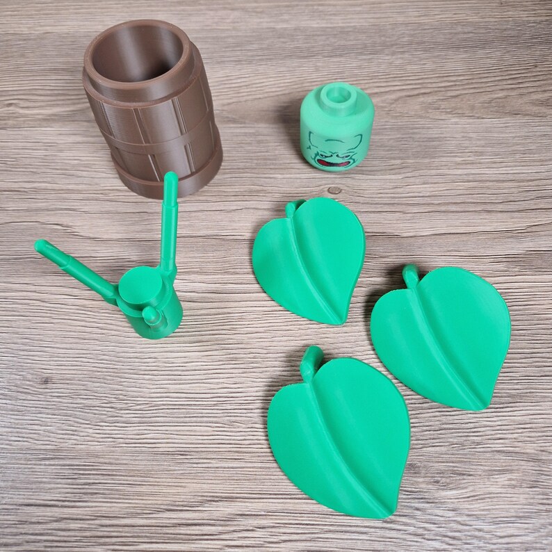 May include: An assortment of 3D-printed items, including a brown barrel-shaped container, a green head-shaped object with a face, a green tool, and three green leaf-shaped objects. The items are arranged on a wooden surface.