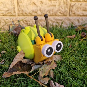 Puede incluir: Un juguete de caracol amarillo y verde, hecho de bloques entrelazados, con ojos grandes y redondos y dos antenas. El caracol est&aacute; sobre una cama de hierba verde y hojas ca&iacute;das, creando una escena l&uacute;dica y colorida.