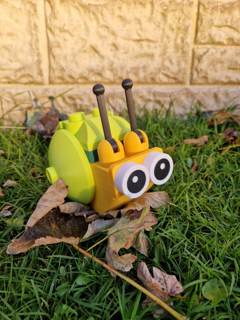 Puede incluir: Un juguete de caracol amarillo y verde, hecho de bloques entrelazados, con ojos grandes y redondos y dos antenas. El caracol est&aacute; sobre una cama de hierba verde y hojas ca&iacute;das, creando una escena l&uacute;dica y colorida.