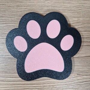 Op de afbeelding: Een zwarte pootafdruk-vormige mat met roze kussentjes. De mat heeft een textuuroppervlak en is ontworpen om op een katten- of hondenpoot te lijken. De achtergrond is een lichtbruin houten oppervlak.