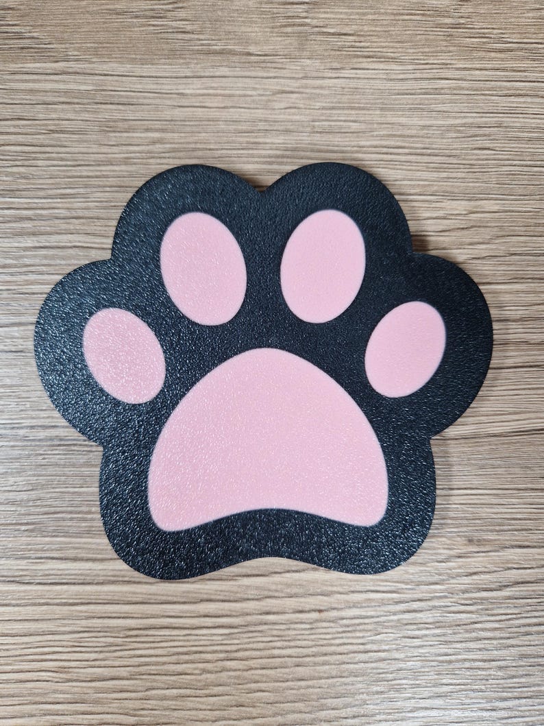 Op de afbeelding: Een zwarte pootafdruk-vormige mat met roze kussentjes. De mat heeft een textuuroppervlak en is ontworpen om op een katten- of hondenpoot te lijken. De achtergrond is een lichtbruin houten oppervlak.