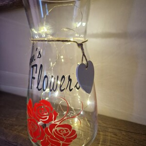 May include: A clear glass vase with a string wrapped around the middle and a small gray heart tag. The vase has a red rose design and the words "Mum's Flowers" written on it. The vase is lit with white fairy lights.