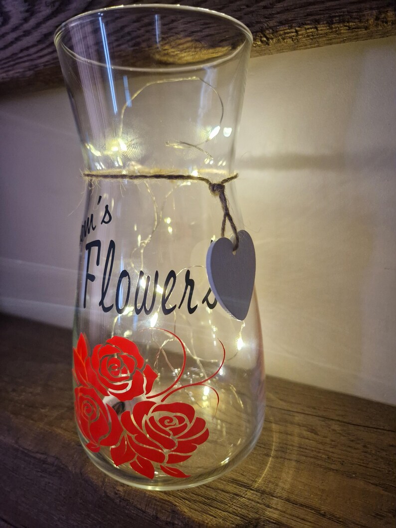 May include: A clear glass vase with a string wrapped around the middle and a small gray heart tag. The vase has a red rose design and the words "Mum's Flowers" written on it. The vase is lit with white fairy lights.