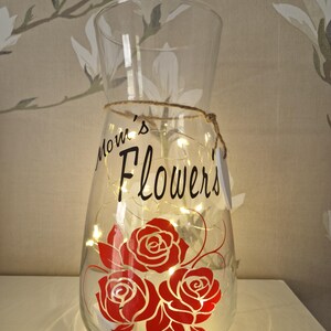 May include: A clear glass vase with a string wrapped around the neck. The vase has a design of red roses and the text "Mom's Flowers" on the front. The vase is lit with white fairy lights.