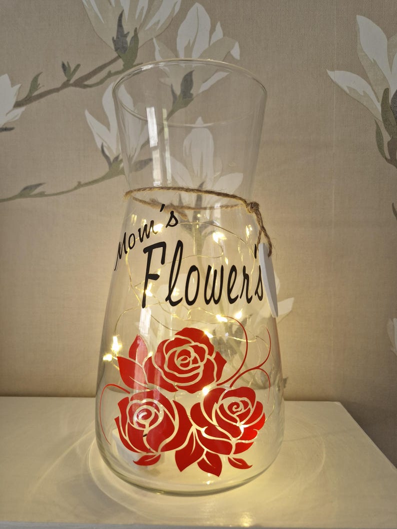 May include: A clear glass vase with a string wrapped around the neck. The vase has a design of red roses and the text "Mom's Flowers" on the front. The vase is lit with white fairy lights.