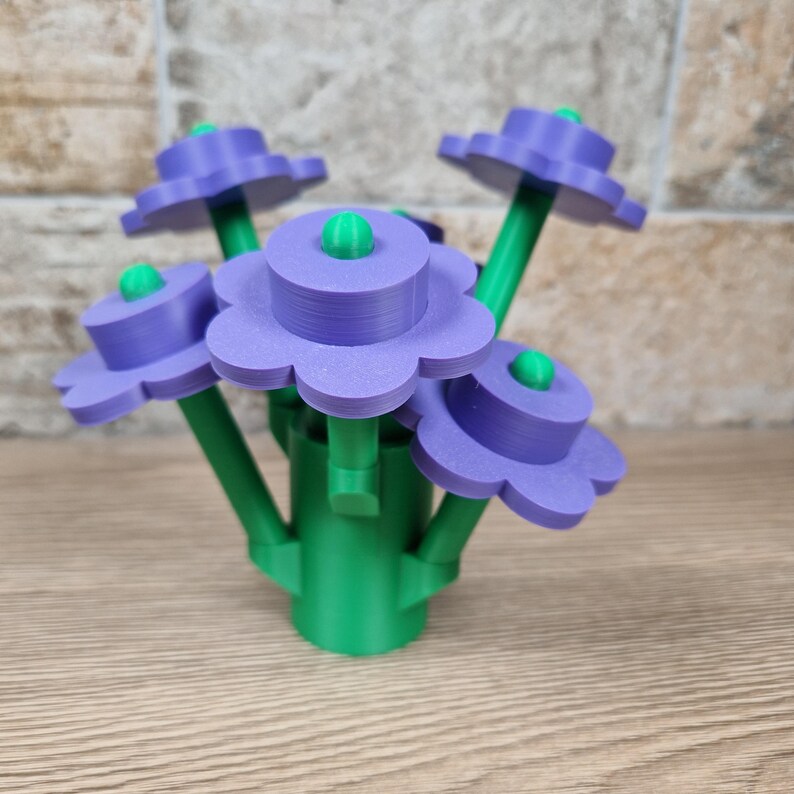 May include: A 3D-printed floral arrangement featuring five stylized flowers. Each flower has a purple, scalloped petal design with a green center. The stems and base are green, creating a vibrant contrast. The arrangement is set against a neutral background.