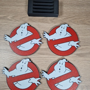 K&ouml;nnte beinhalten: Vier runde Untersetzer mit dem Ghostbusters-Logo: ein wei&szlig;er Geist in einem roten Kreis mit einer diagonalen Linie. Ein schwarzer, rechteckiger Untersetzerhalter ist ebenfalls vorhanden. Die Untersetzer sind aus einem holz&auml;hnlichen Material.