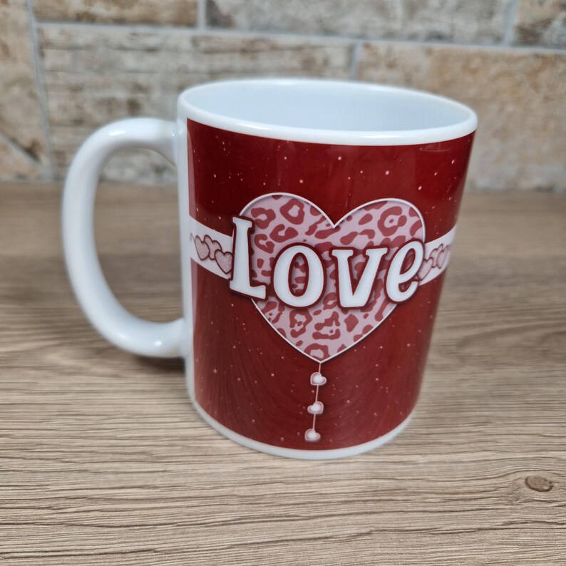 May include: A white ceramic mug with a red background and a heart-shaped design. The heart features a leopard print pattern and the word "LOVE" in white. The mug has a white handle and a white interior.