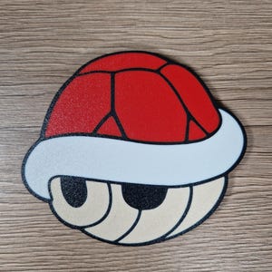 K&ouml;nnte beinhalten: Eine rote, wei&szlig;e und beige Cartoon-Schildkr&ouml;tenpanzer. Die Schale hat eine rote Oberseite mit schwarzen Linien, einen wei&szlig;en Rand und beige Augen und Mund. Das Design erinnert an einen Videospielcharakter.