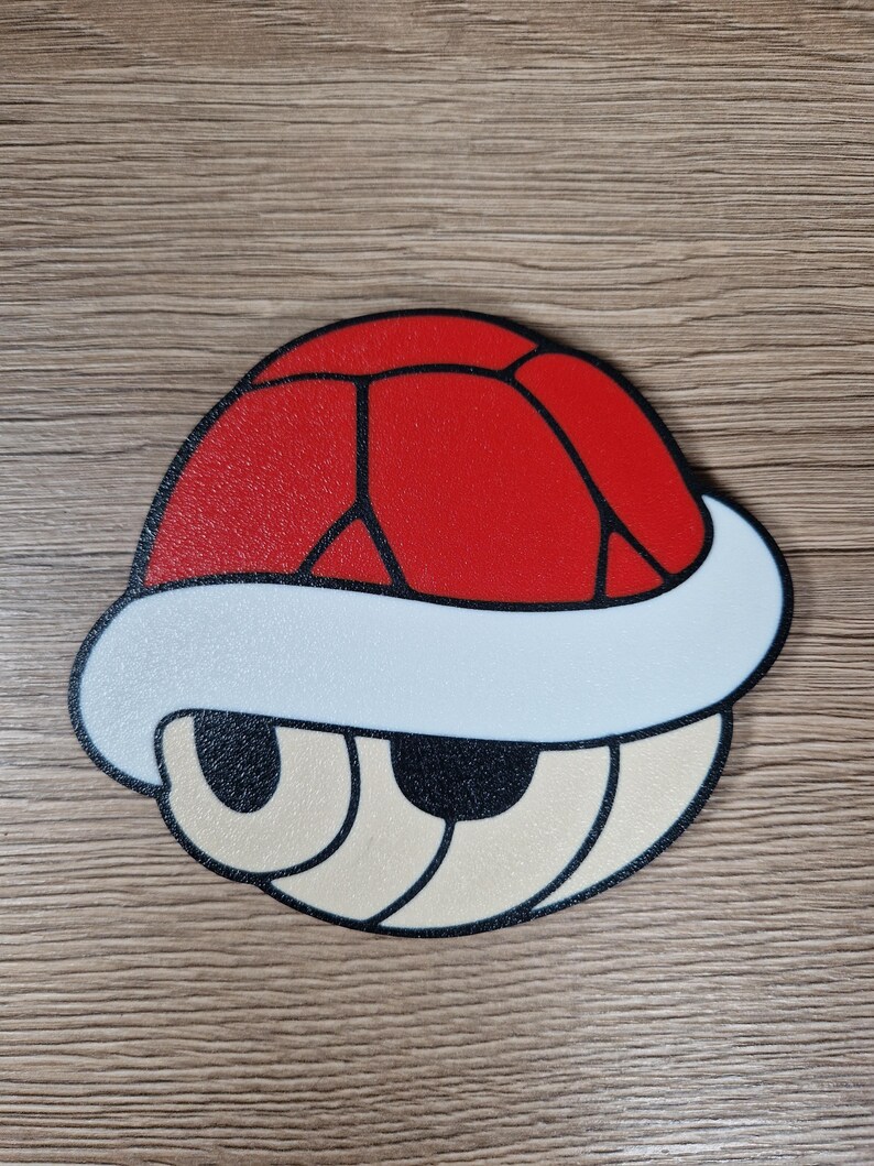 K&ouml;nnte beinhalten: Eine rote, wei&szlig;e und beige Cartoon-Schildkr&ouml;tenpanzer. Die Schale hat eine rote Oberseite mit schwarzen Linien, einen wei&szlig;en Rand und beige Augen und Mund. Das Design erinnert an einen Videospielcharakter.