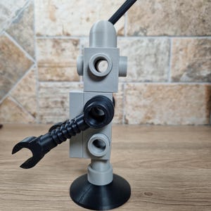 May include: A gray and black robot-like sculpture with a rounded head, circular openings, and a wrench-shaped arm. The sculpture has a black antenna and a circular base. The robot is made of plastic and sits on a wooden surface.