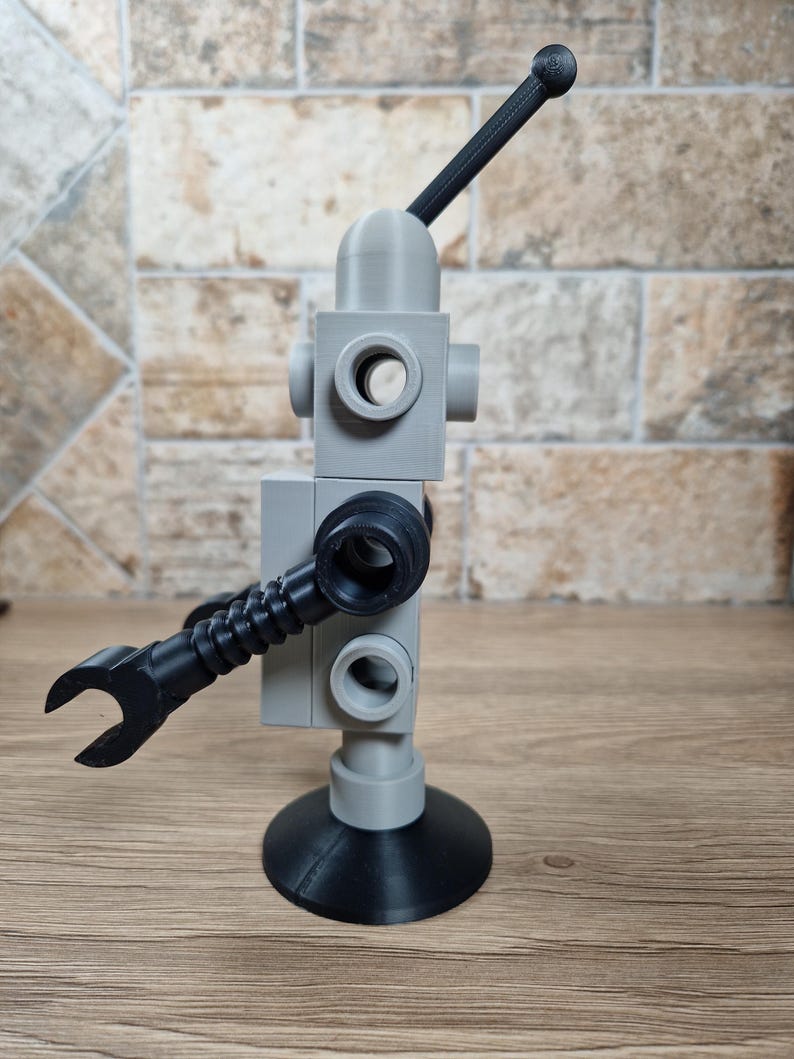 May include: A gray and black robot-like sculpture with a rounded head, circular openings, and a wrench-shaped arm. The sculpture has a black antenna and a circular base. The robot is made of plastic and sits on a wooden surface.