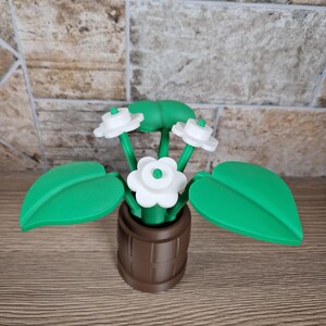 May include: A decorative, artificial flower arrangement featuring white flowers with green centers and stems, and large green leaves. The flowers are in a brown barrel-shaped pot. The arrangement is made of plastic.