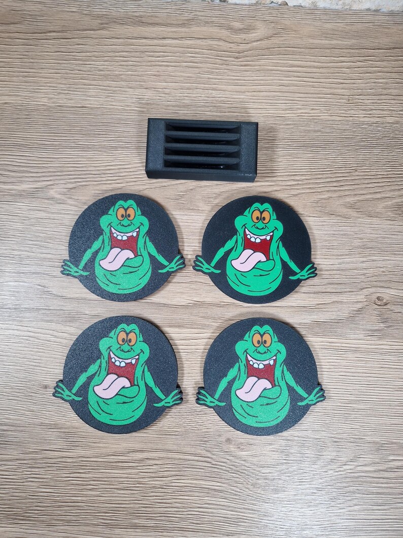 May include: Four black coasters with a green cartoon character with a long tongue and a black rectangular object. The character is green with a pink tongue and a white mouth. The coasters are round.