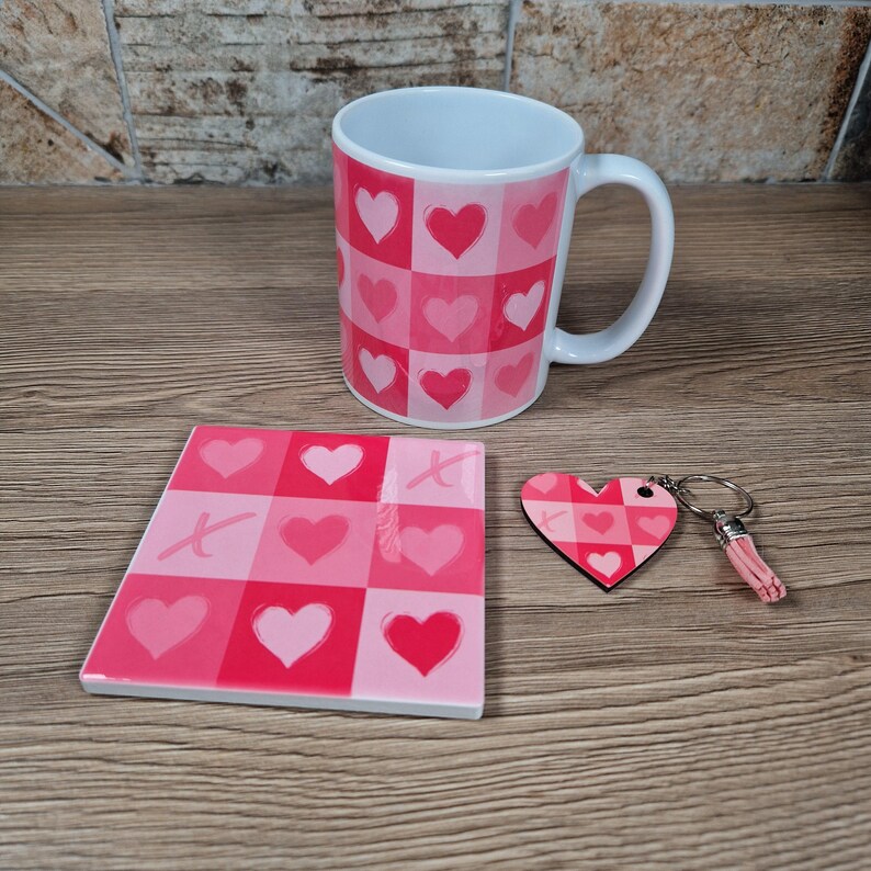 May include: A white ceramic mug, coaster, and heart-shaped keychain with a pink heart pattern. The mug and keychain have a grid of hearts in various shades of pink. The coaster has a tic-tac-toe design with hearts and Xs.