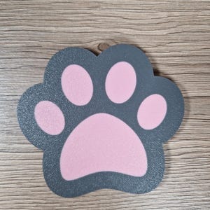 Op de afbeelding: Een grijze pootafdruk-vormige onderzetter met een roze pootkussenontwerp. De onderzetter is gemaakt van een duurzaam materiaal en is ontworpen om oppervlakken te beschermen tegen krassen en waterringen. Het pootafdrukontwerp is een schattige en speelse toevoeging.