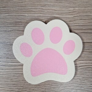 Op de afbeelding: Een pootvormig decoratief item met een witte achtergrond en roze voetzolen. De pootafdruk heeft een glittertextuur. Het item is waarschijnlijk gemaakt van hout of een vergelijkbaar materiaal. Het ontwerp is eenvoudig en schattig, geschikt voor dierenliefhebbers.