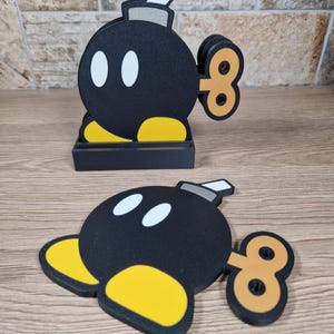 May include: Black, yellow, and white cartoon bomb-shaped coasters. The coasters have a wind-up key design and are in a black holder. One coaster is lying flat on a wooden surface, and another is standing upright.