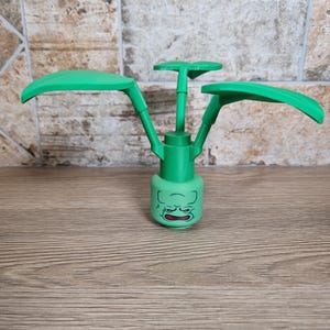 May include: A green plastic toy plant with a face on the base and three large leaves. The toy is made of green plastic and has a cartoon-like appearance. The base has a face with a grumpy expression.