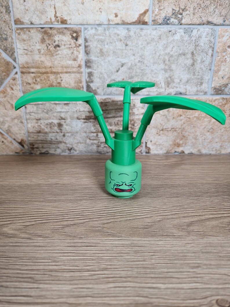 May include: A green plastic toy plant with a face on the base and three large leaves. The toy is made of green plastic and has a cartoon-like appearance. The base has a face with a grumpy expression.