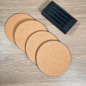 May include: Four round cork coasters with black edges are arranged on a light wood-grain surface. A black rectangular holder with vertical slots sits nearby. The coasters are a natural tan color.