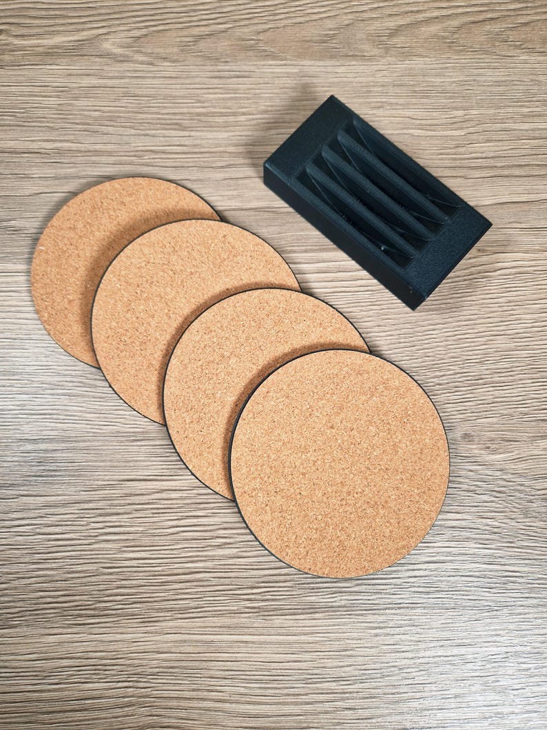 May include: Four round cork coasters with black edges are arranged on a light wood-grain surface. A black rectangular holder with vertical slots sits nearby. The coasters are a natural tan color.
